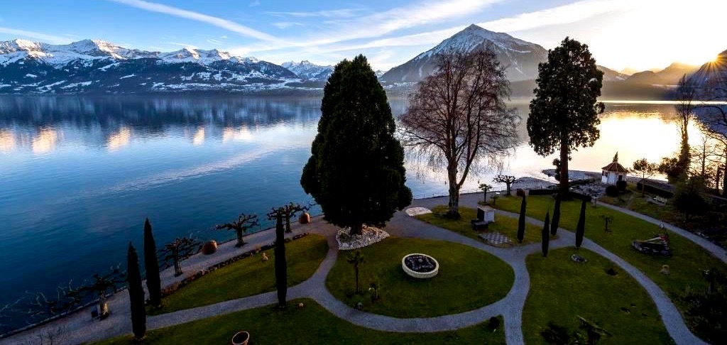 Ansicht vom grossen Park des Parkhotels Gunten mit Thunersee und Berge im Schnee bei klarem sonnigen Wetter