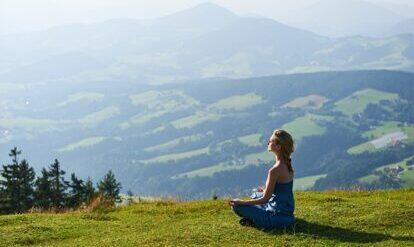 Frau im Yogasitz auf einem Wiesenhügel mit Berglandschaft