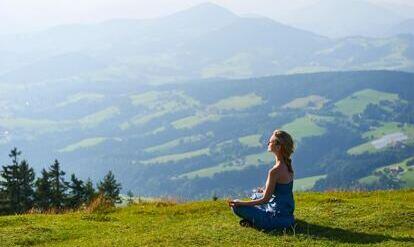Frau im Yogasitz auf einem Wiesenhügel mit Berglandschaft