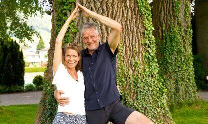 Yogalehrer Ernst und Yogalehrerin Sibylle in der Yogastellung Baum, vor einem Baum auf der Wiese 