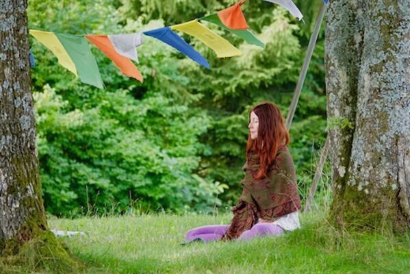 Claudia Eva Reinig sitzt im Yogasitz in Meditation unter einem Baum, tibetische Gebetsfahnen hängen