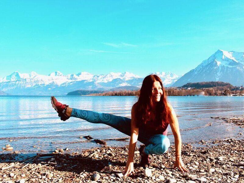Claudia Reinig in einer Yoga Position Hüftöffner, Hocke mit rechtem Bein nach aussen gestreckt, im Hintergrund Thunersee und Alpen