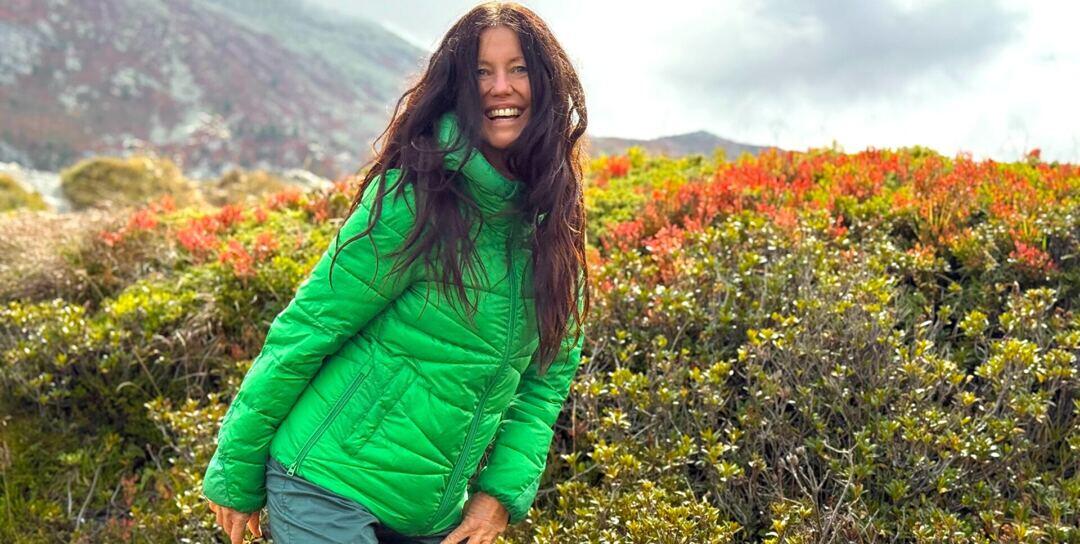 Yogalehrerin Claudia Eva Reinig im Herbst mit grüner Daunenjacke vor roten Alpenrosen, im Hintergrund Berge