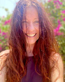 Claudia Eva Reinig - Portrait - lachend in der Sonne mit rosafarbenen Blumen im Hintergrund